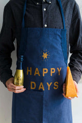 Man holding champagne and cheese, wearing 'Happy Days' apron